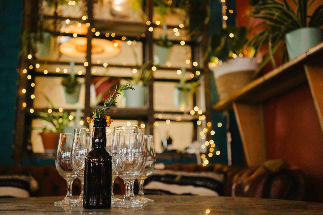 Cozy cafe setting with wine glasses, a bottle, plants, and string lights.